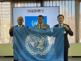 国連旗の贈呈式で、CIFALジャパンの代表者と国連ユニタール人々・社会包摂部長のアレックス・メヒア氏が国連旗を掲げる様子。